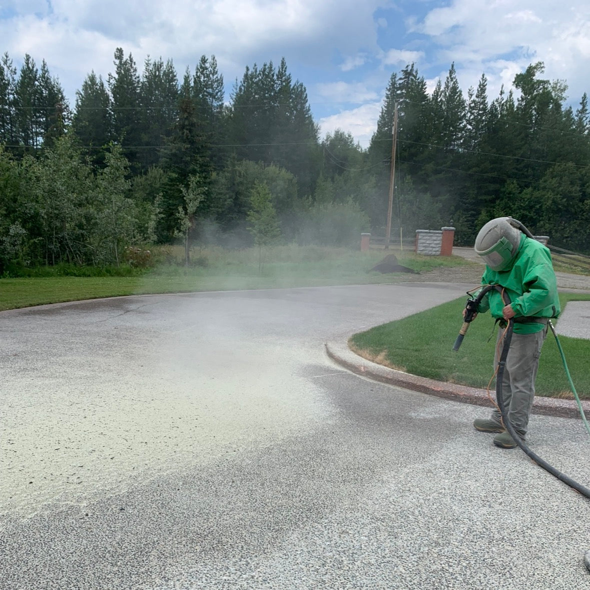 Person applying sandblasting to a road using a spray gun, with trees and a clear sky in the background.